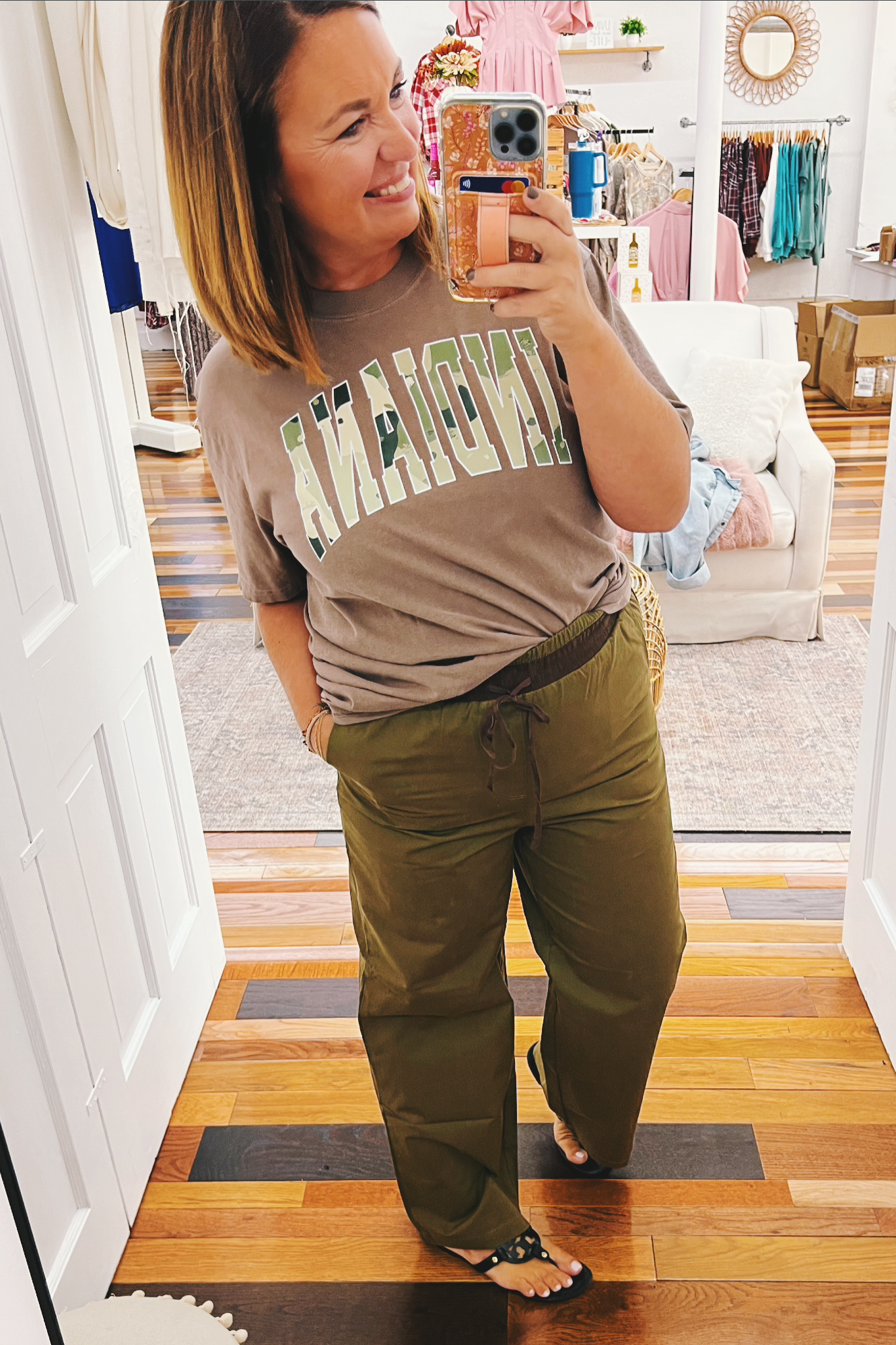 Woman taking a mirror selfie wearing a graphic t-shirt and green pants in a store.
