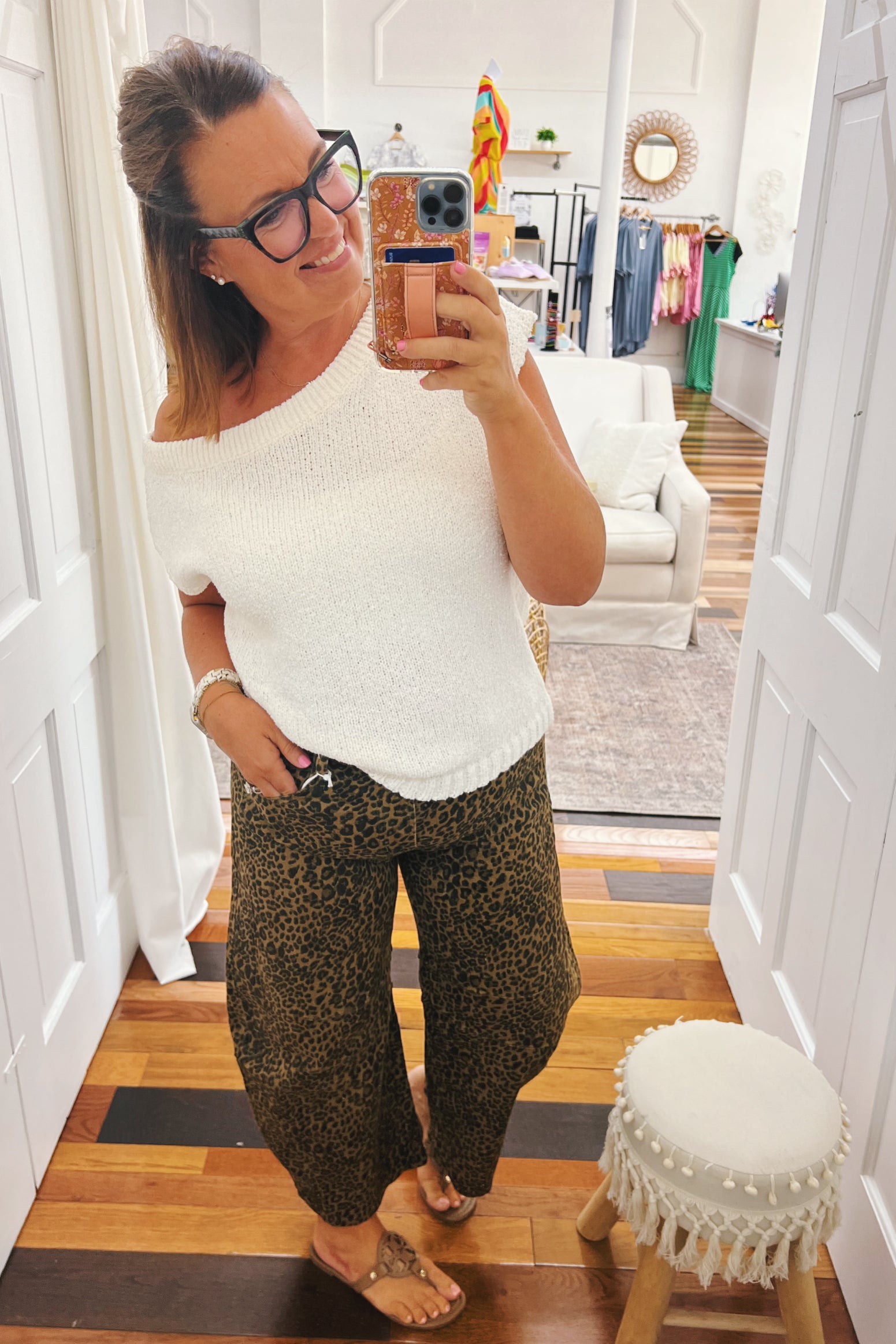 Woman taking a mirror selfie wearing a white top and leopard print pants in a room with wooden flooring.